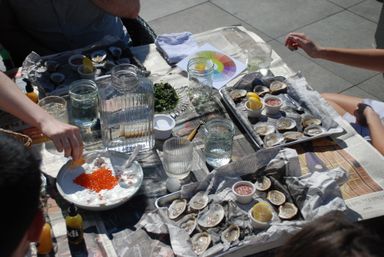 Sunlit outdoor patio table with freshly shucked oysters on newspaper-lined trays, lemon wedges and dipping sauces, mason jars of water and a bowl of bright orange fish roe being served — casual seafood gathering.
