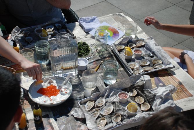 Sunlit outdoor patio table with freshly shucked oysters on newspaper-lined trays, lemon wedges and dipping sauces, mason jars of water and a bowl of bright orange fish roe being served — casual seafood gathering.