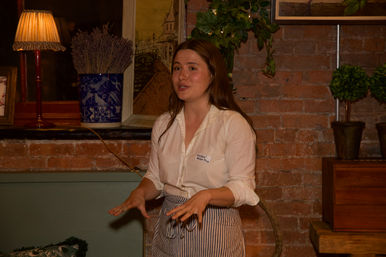 Young woman in a white shirt and striped apron speaking and gesturing in a cozy brick-walled room lit by a warm lamp, with potted plants and a blue-and-white vase in the background.