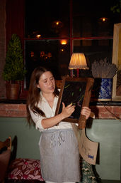 Server in a striped apron pointing to a small chalkboard menu in a cozy brick-walled restaurant interior by a window, warm table lamp, potted topiary and a blue vase of dried lavender visible.