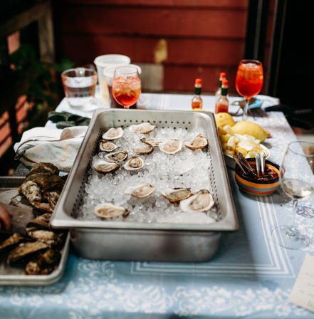 Raw oysters on ice in a stainless tray with lemon wedges, hot sauce and bright cocktails on an outdoor patio table