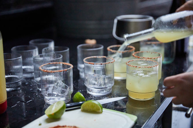 Pouring pale lime cocktails into ribbed glasses with chili-rimmed edges, ice-filled glasses, lime wedges and a knife on a glossy bar countertop — casual cocktail prep scene.