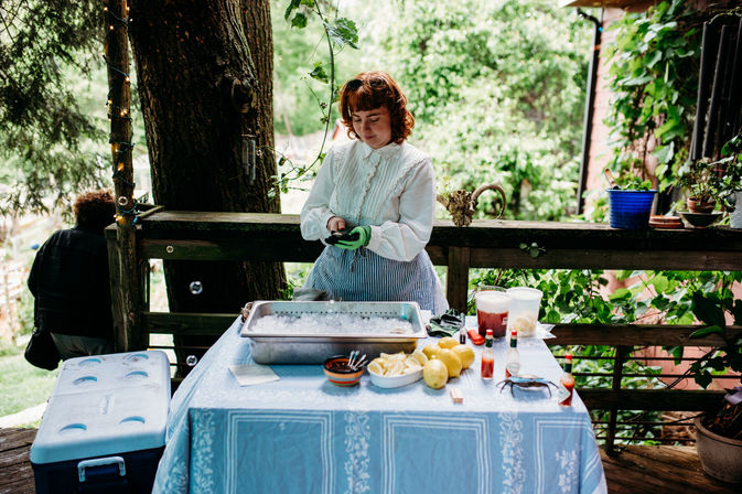 Person in a white blouse and striped apron wearing green gloves, prepping food at an outdoor garden deck table with an ice-filled metal tray, lemons, condiments, cooler and potted plants nearby.
