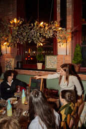 Server presenting a tasting-menu chalkboard to diners at a cozy brick-walled downtown restaurant with hanging floral lights and table wine.