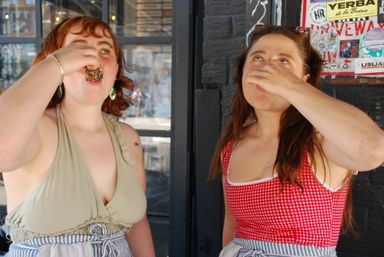 Two women in summer tops and aprons stand outside a storefront, tilting their heads back and playfully taking shots from small glasses.