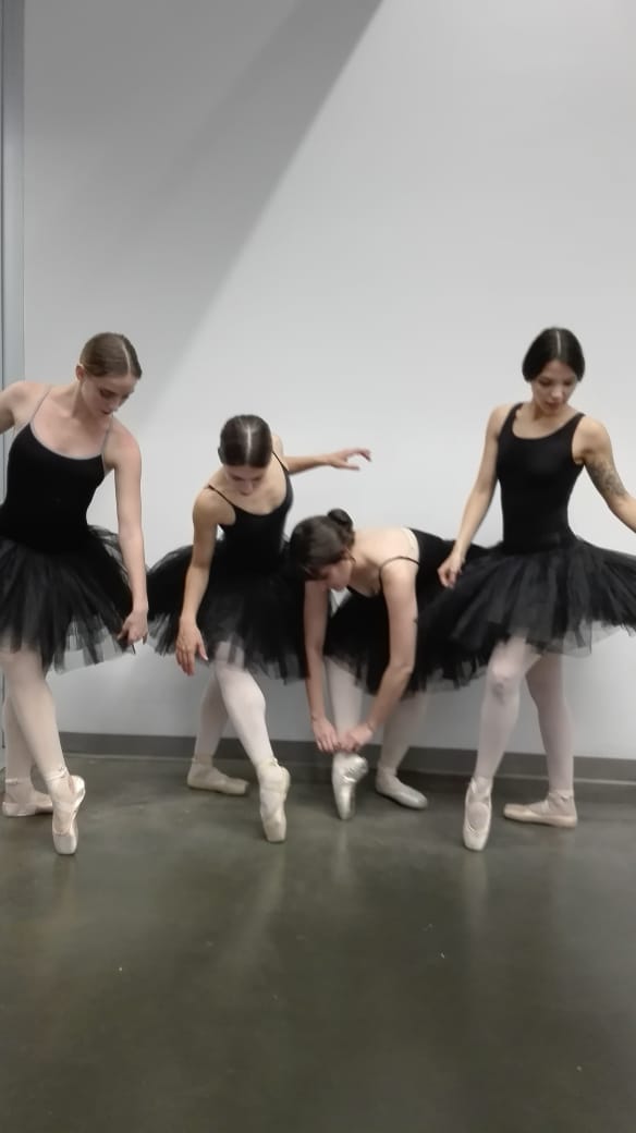 Four ballet dancers in black tutus and pink pointe shoes adjusting footwear and posing against a white dance studio wall on a gray floor.