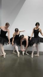 Four ballet dancers in black tutus and pink pointe shoes adjusting footwear and posing against a white dance studio wall on a gray floor.