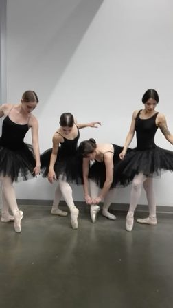 Four ballet dancers in black tutus and pink pointe shoes adjusting footwear and posing against a white dance studio wall on a gray floor.