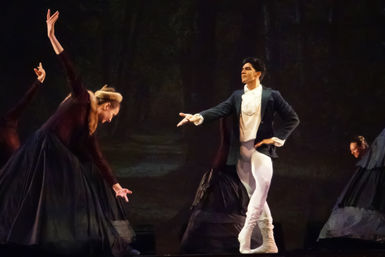 Classical ballet on a theater stage: male dancer in tailcoat and white tights strikes a dramatic pose toward bowing female dancers in dark period skirts against a painted forest backdrop.