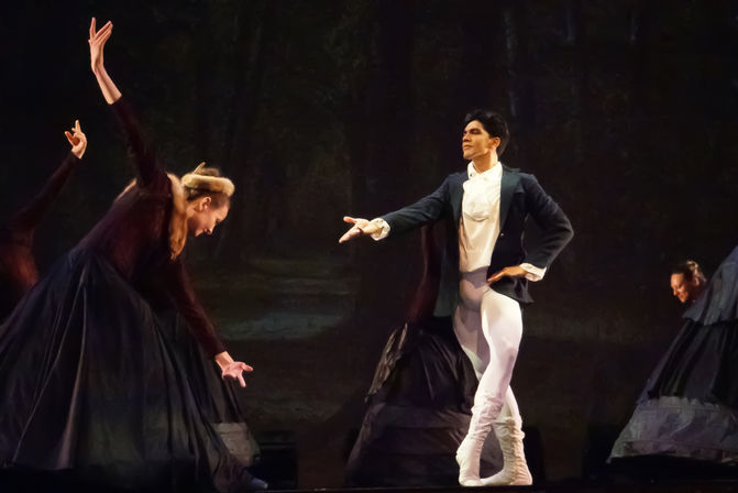 Classical ballet on a theater stage: male dancer in tailcoat and white tights strikes a dramatic pose toward bowing female dancers in dark period skirts against a painted forest backdrop.