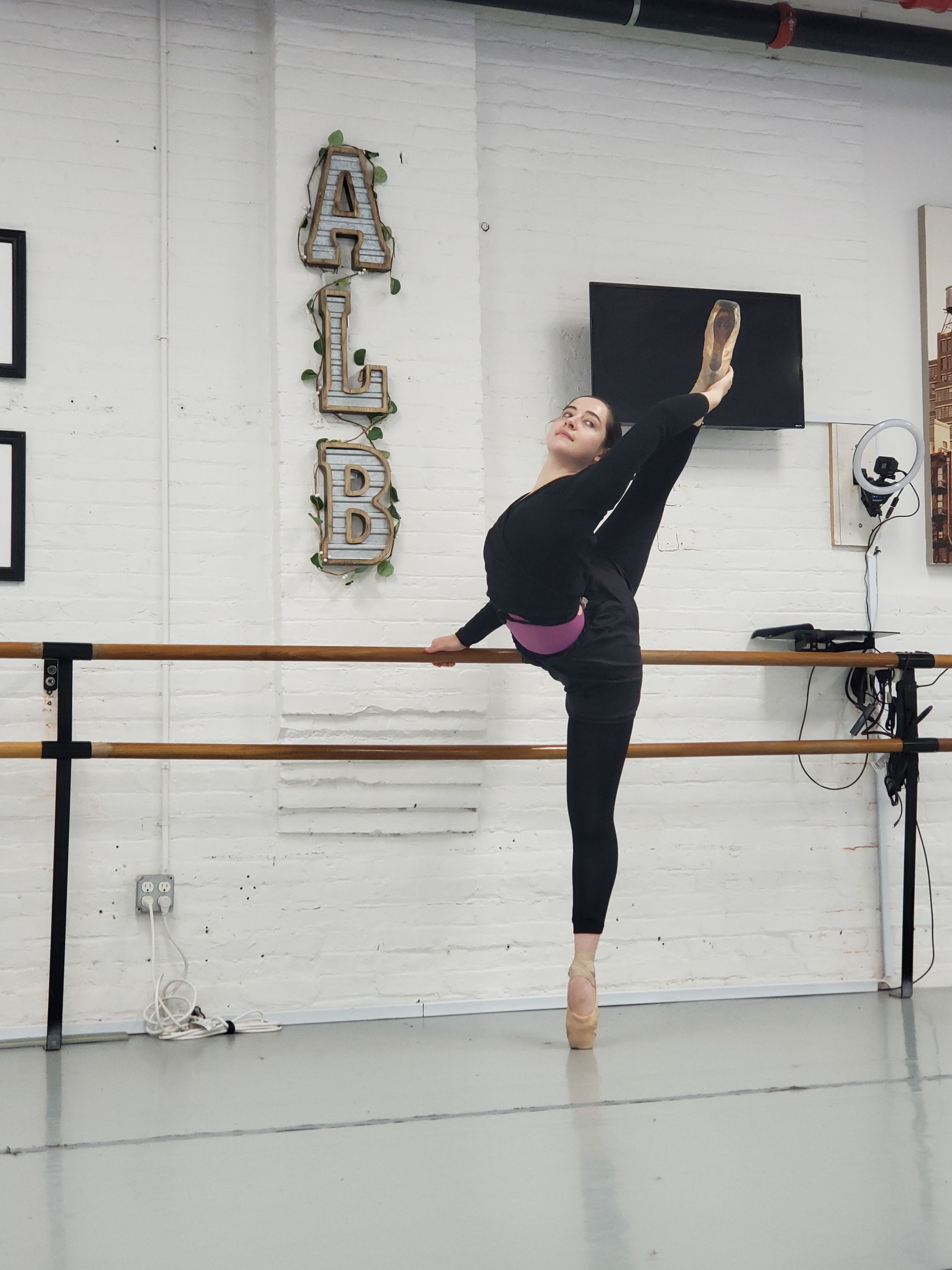 Ballet dancer in black practicewear on pointe performing a high needle-style leg extension at a wooden barre in a white-brick dance studio