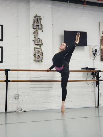 Ballet dancer in black practicewear on pointe performing a high needle-style leg extension at a wooden barre in a white-brick dance studio