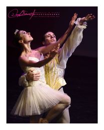 Two ballet dancers performing a graceful pas de deux on a dark stage — female in a white tutu and tiara lifted by a male in a gold brocade vest, both reaching upward.