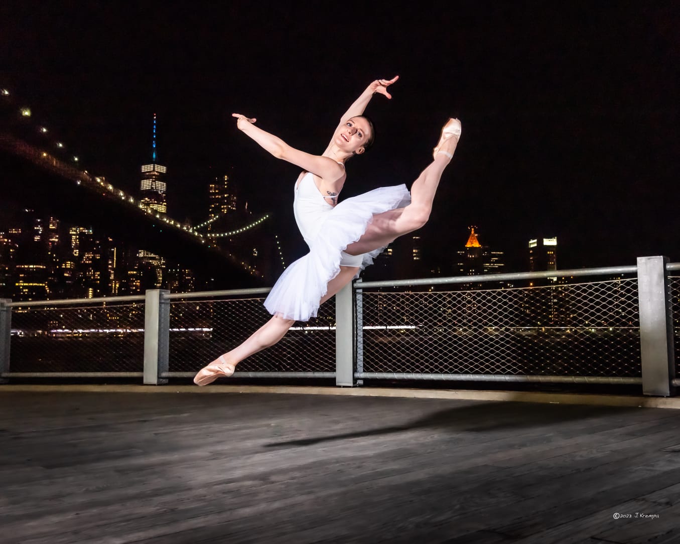 Ballerina in a white tutu leaping en pointe on a waterfront pier at night with the illuminated New York City skyline and suspension bridge glowing behind her