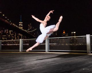 Ballerina in a white tutu leaping en pointe on a waterfront pier at night with the illuminated New York City skyline and suspension bridge glowing behind her