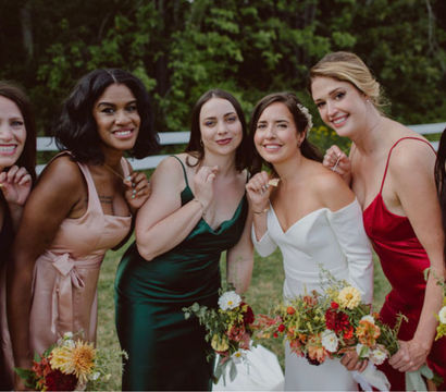 Outdoor wedding portrait of the bride and five bridesmaids posing by a white fence in a green field, smiling and holding colorful autumn bouquets; dresses in white, emerald, red, blush, navy and orange satin for a rustic garden celebration.
