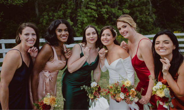 Outdoor wedding portrait of the bride and five bridesmaids posing by a white fence in a green field, smiling and holding colorful autumn bouquets; dresses in white, emerald, red, blush, navy and orange satin for a rustic garden celebration.