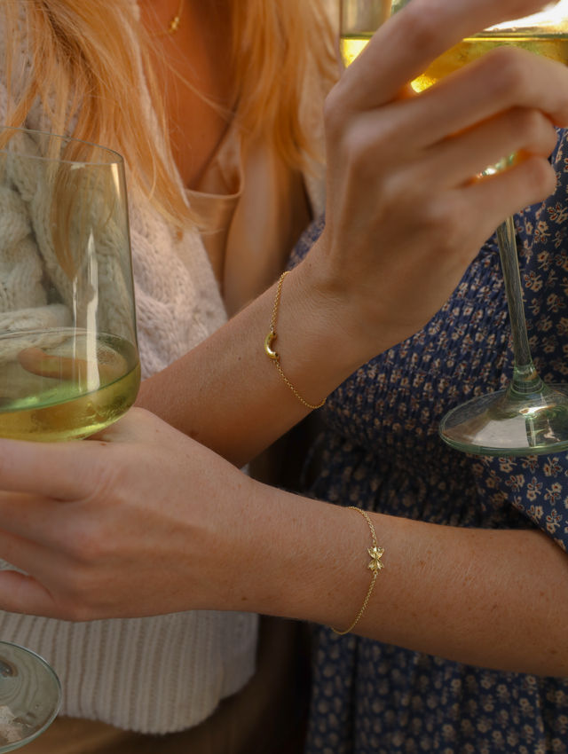 Sunlit close-up of two people holding white wine glasses, highlighting delicate gold charm bracelets on their wrists against cozy knit and floral dress fabrics.