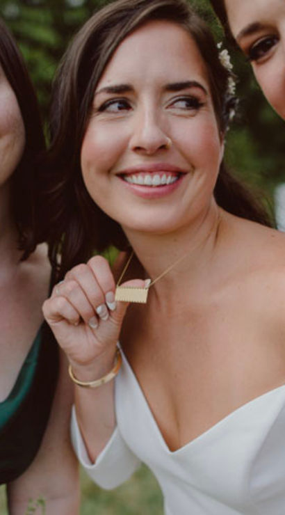 Three smiling women at an outdoor wedding — bride in an off-shoulder white gown and two bridesmaids in green and red satin showing necklaces