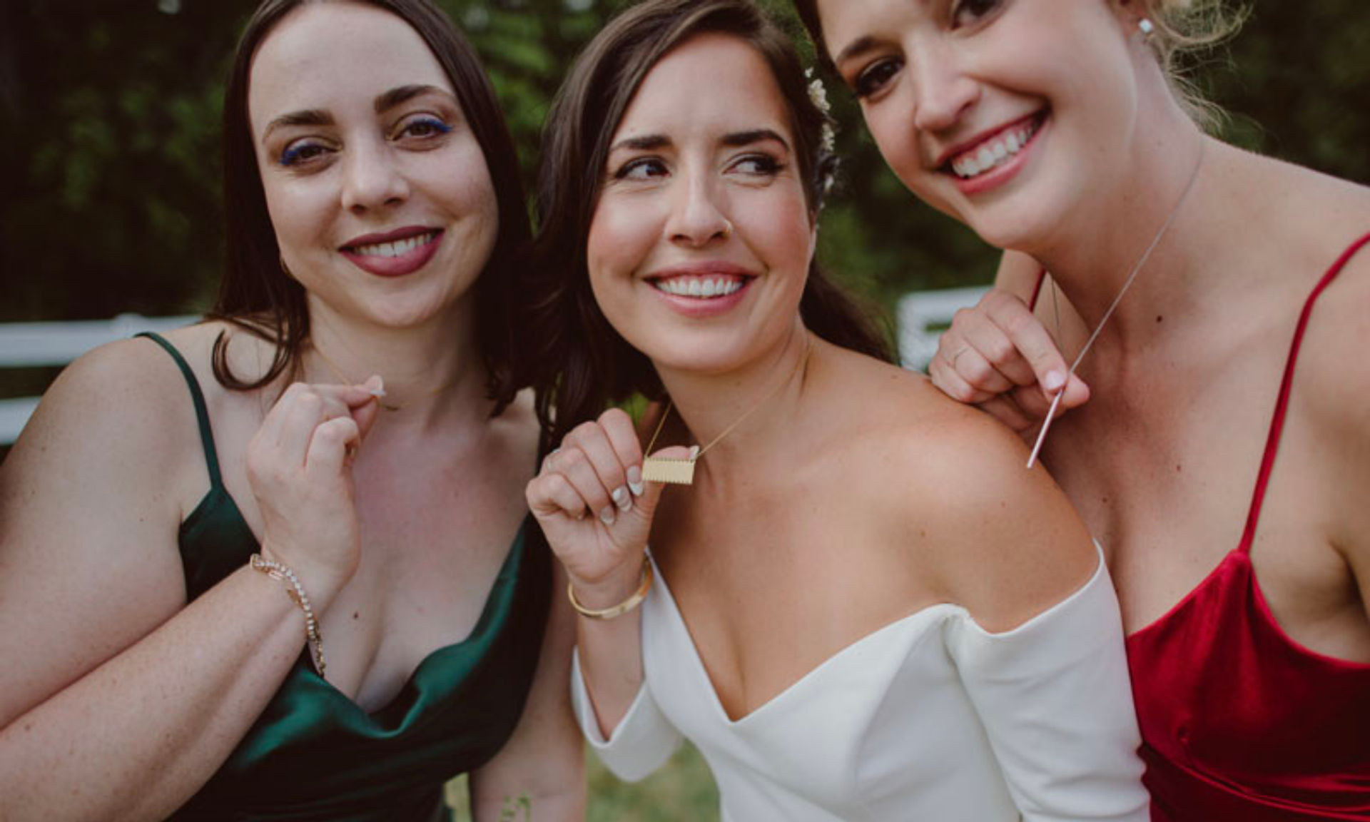 Three smiling women at an outdoor wedding — bride in an off-shoulder white gown and two bridesmaids in green and red satin showing necklaces