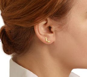 Close-up profile of a redhead wearing a small gold crescent stud earring — minimalist jewelry detail on the ear against a white background.