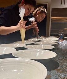 Two chefs piping sauces from pastry bags into a line of white bowls on a professional kitchen counter — close-up of restaurant plating prep.