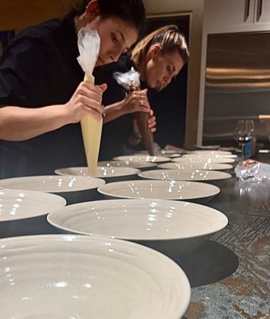 Two chefs piping sauces from pastry bags into a line of white bowls on a professional kitchen counter — close-up of restaurant plating prep.