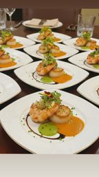 Seared scallop and shrimp appetizers on white plates, each plated with bright orange and green purées, balsamic drizzle, microgreens and an edible flower, arranged on a dark wood table in a fine-dining setting.
