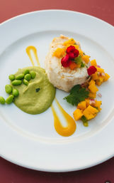 Gourmet plated white fish roulade with mango salsa, edamame purée and mango coulis, garnished with cilantro and a red edible flower on a white plate over a red table.