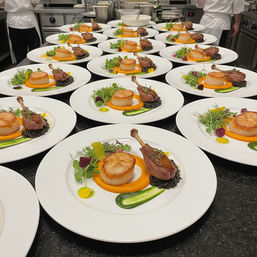 Row of fine-dining plates in a restaurant kitchen: seared scallop and confit duck leg on carrot purée with black lentils, green herb smear and microgreens, ready for tasting-menu service.