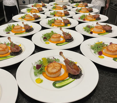 Row of fine-dining plates in a restaurant kitchen: seared scallop and confit duck leg on carrot purée with black lentils, green herb smear and microgreens, ready for tasting-menu service.
