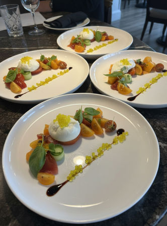 Artfully plated burrata salad with heirloom cherry tomatoes, cucumber ribbons, fresh basil, balsamic drizzle and bright yellow molecular pearls on white plates in a restaurant setting.