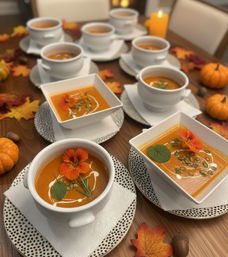 Autumn dining table with white bowls of creamy butternut squash soup garnished with orange edible flowers, pumpkin seeds, cream swirls and herbs, surrounded by mini pumpkins and fall leaves.