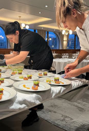 Two chefs carefully plating colorful bite-sized gourmet appetizers on white china in an elegant fine-dining room with arched windows and city skyline at dusk.