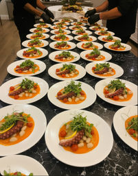 Rows of white plates on a black marble counter for a catered event, each with a grilled octopus tentacle over red pepper purée, white beans, cherry tomatoes, lemon and microgreens as chefs in gloves finish plating.