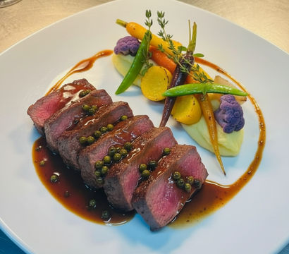 Gourmet sliced medium-rare steak drizzled with green peppercorn jus, served with creamy mashed potatoes, heirloom carrots, purple cauliflower and snap peas on a white plate