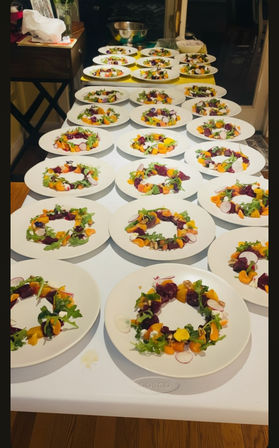 Long table lined with dozens of white plates holding colorful salad rings — arugula, sliced radishes, roasted beets, orange segments, nuts and dollops of cheese — plated for a catered dinner or large dinner party.