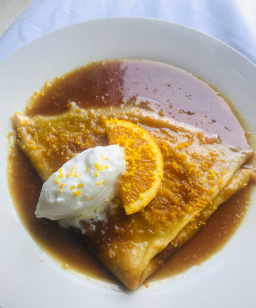 Folded crepe on a white plate drenched in orange caramel sauce, topped with a dollop of whipped cream, an orange slice and bright orange zest — citrus dessert or brunch treat.