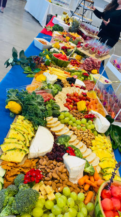 Vibrant catered grazing table at an indoor event with a long charcuterie spread of cheeses, crackers, nuts, sliced pineapple, grapes, watermelon, cherry tomatoes, broccoli, baby carrots and mini dessert cups on a blue tablecloth.