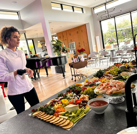 Chef in a white jacket stands by a party-ready charcuterie and shrimp cocktail spread on a kitchen island in a sunlit modern open-plan home with piano and chairs set for guests.