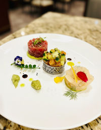 Fine-dining seafood trio on a white plate: tuna tartare cylinder on avocado, diced cucumber‑salmon medley in an ornate ring topped with gold leaf, and scallop carpaccio garnished with edible petal, dill, wasabi dollops, balsamic dots and microgreens.