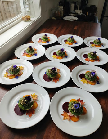 Rows of white plates on a wooden countertop by a window, each with an autumn-inspired gourmet appetizer — a green herb-topped round sprinkled with seeds, colorful beet and golden slices, radish accents, maple-shaped garnish and a purple edible flower.