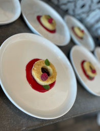 Ring-shaped mini cake on bright raspberry coulis, garnished with a fresh raspberry and mint leaf on a white plate, with several identical plated desserts blurred in the background.