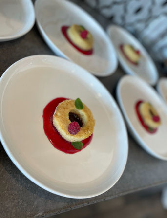 Ring-shaped mini cake on bright raspberry coulis, garnished with a fresh raspberry and mint leaf on a white plate, with several identical plated desserts blurred in the background.