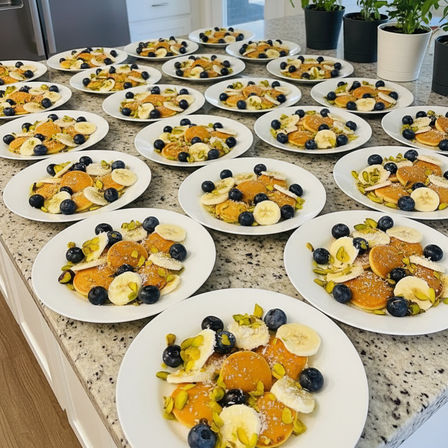 Rows of white plates on a granite kitchen island, each with mini pancakes topped with banana slices, blueberries, pistachios and shredded coconut — ready-to-serve brunch spread