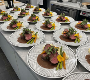 Rows of fine-dining plates on a marble pass in a professional kitchen featuring slices of medium-rare beef medallions with rich brown sauce, mashed potatoes, asparagus, baby carrots and edible flowers, lined up and ready for service.