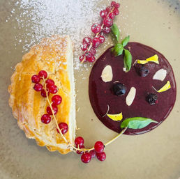 Top-down view of a plated gourmet dessert: flaky golden half-moon pastry dusted with powdered sugar, glossy dark berry coulis with blueberries and almond slices, edible yellow petals and basil, garnished with red currant sprig.