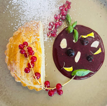 Top-down view of a plated gourmet dessert: flaky golden half-moon pastry dusted with powdered sugar, glossy dark berry coulis with blueberries and almond slices, edible yellow petals and basil, garnished with red currant sprig.