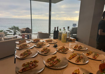 Oceanfront kitchen counter in a beachfront home with dozens of plated open-faced appetizers and bottles lined up, sunset over the sea visible through floor-to-ceiling windows.