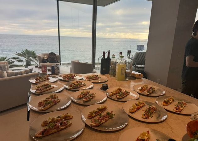 Oceanfront kitchen counter in a beachfront home with dozens of plated open-faced appetizers and bottles lined up, sunset over the sea visible through floor-to-ceiling windows.